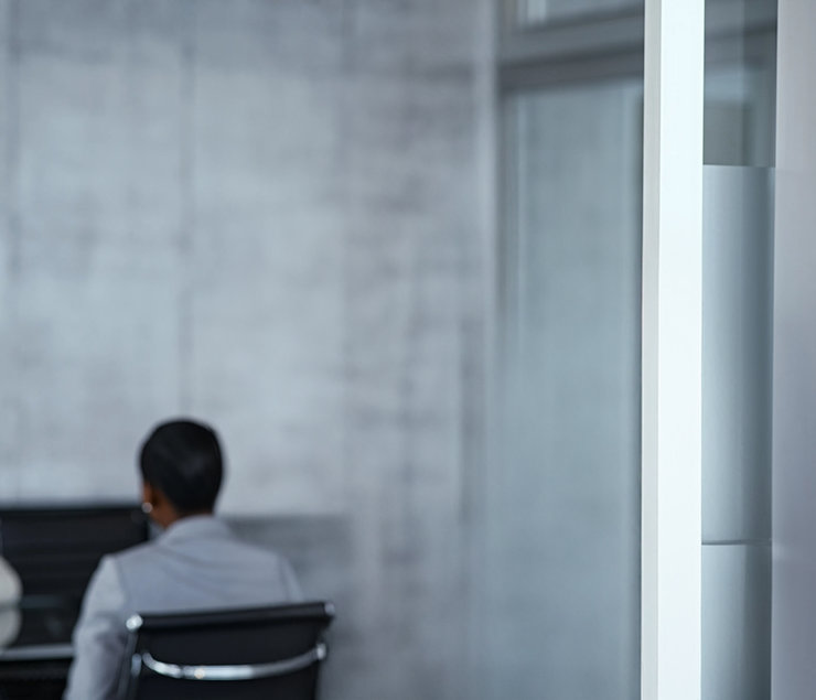 Besprechung im modernen Büro Zwei Personen sitzen in einem modernen Konferenzraum. Eine Person ist von hinten zu sehen, mit kurzen Haaren und einem grauen Anzug. Die andere Person ist im Hintergrund weniger sichtbar. Der Raum hat eine minimalistische Gestaltung mit Betonwänden und einem großen Fenster.