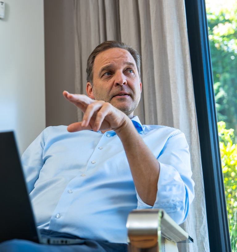 Geschäftsmann mit Laptop im Gespräch Ein Mann sitzt auf einem Stuhl und zeigt mit seiner Hand. Vor ihm steht ein Laptop. Er trägt ein hellblaues Hemd und hat einen nachdenklichen Gesichtsausdruck. Im Hintergrund sind Vorhänge und ein Fenster mit Blick ins Freie.