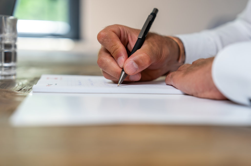 Hand schreibt in Notizbuch Eine Hand hält einen Stift und schreibt in ein Notizbuch, das auf einem Holztisch liegt. Im Hintergrund ist ein Wasserglas und ein Fenster zu sehen.