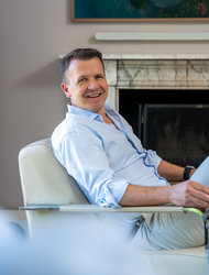 Lächelnder Mann im Wohnzimmer Ein lächelnder Mann sitzt entspannt in einem modernen Sessel. Er trägt ein hellblaues Hemd und hat kurze, dunkle Haare. Im Hintergrund ist ein Kamin und ein Gemälde sichtbar. Der Mann strahlt Freundlichkeit und Gelassenheit aus.